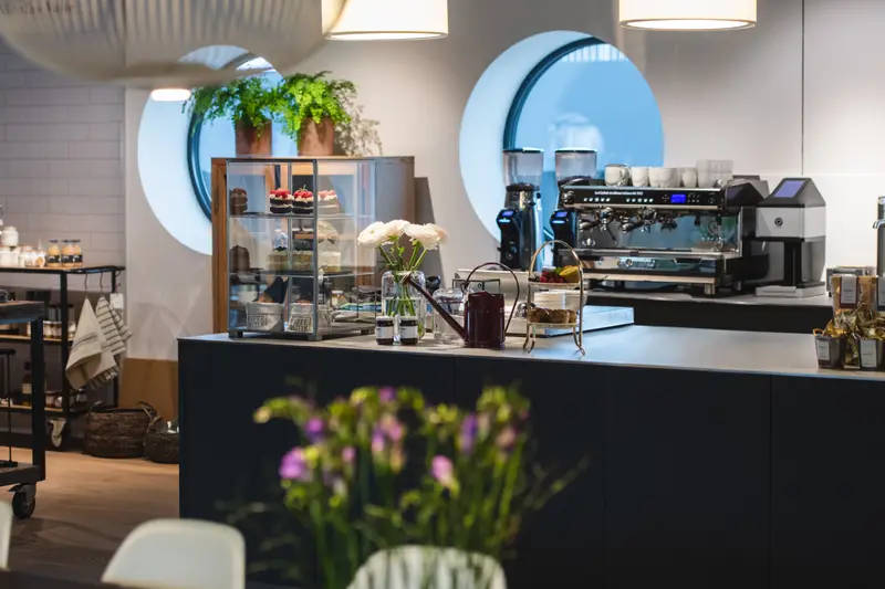 Theke mit Kaffeemaschine und Küchlein-Vitrine vor den runden Fenstern